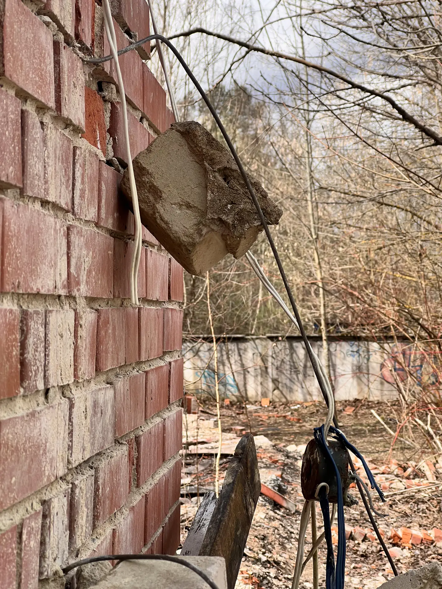 Broken masonry and hanging cables beside a brick wall and young trees.